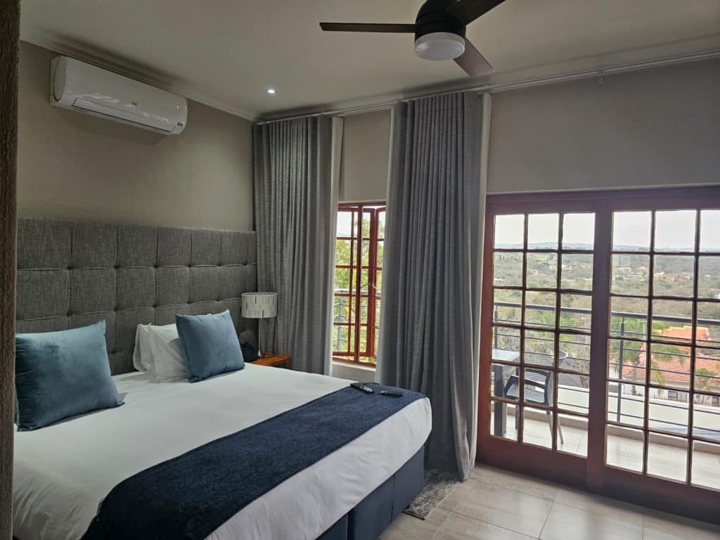 A modern bedroom with a large bed featuring blue pillows and a dark blue throw. Gray curtains cover large windows and a door leading to a balcony with a view. A ceiling fan and air conditioning unit are visible on the ceiling and wall.