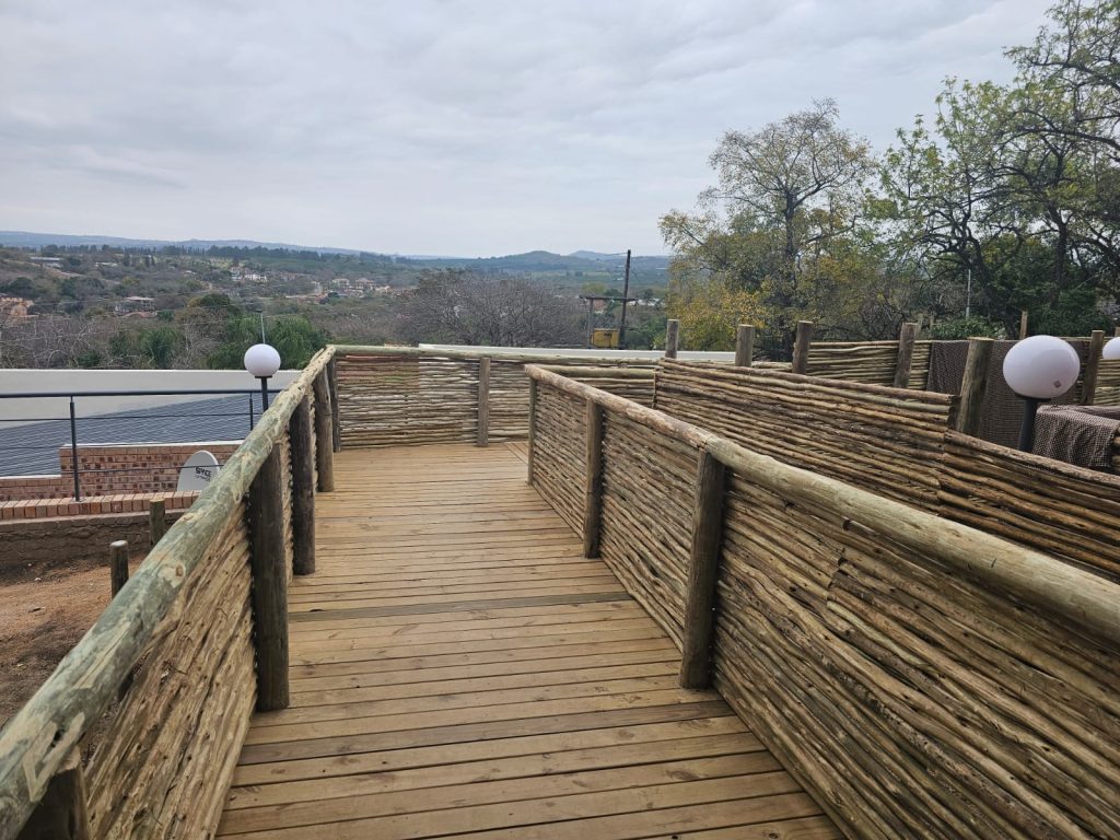 A wooden pathway with rustic log railings leads to a scenic view of a distant landscape. The sky is overcast, and there are trees and hills in the background. Round white lights are mounted along the path.