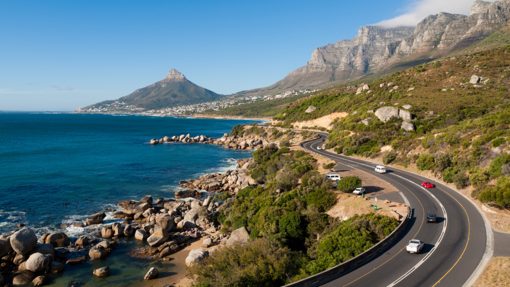 A winding coastal road hugs the shoreline, with cars driving along. The landscape features rocky beaches, blue ocean waters, and rugged mountains under a clear blue sky. Lush greenery lines the road, with a distinctive mountain peak in the background.