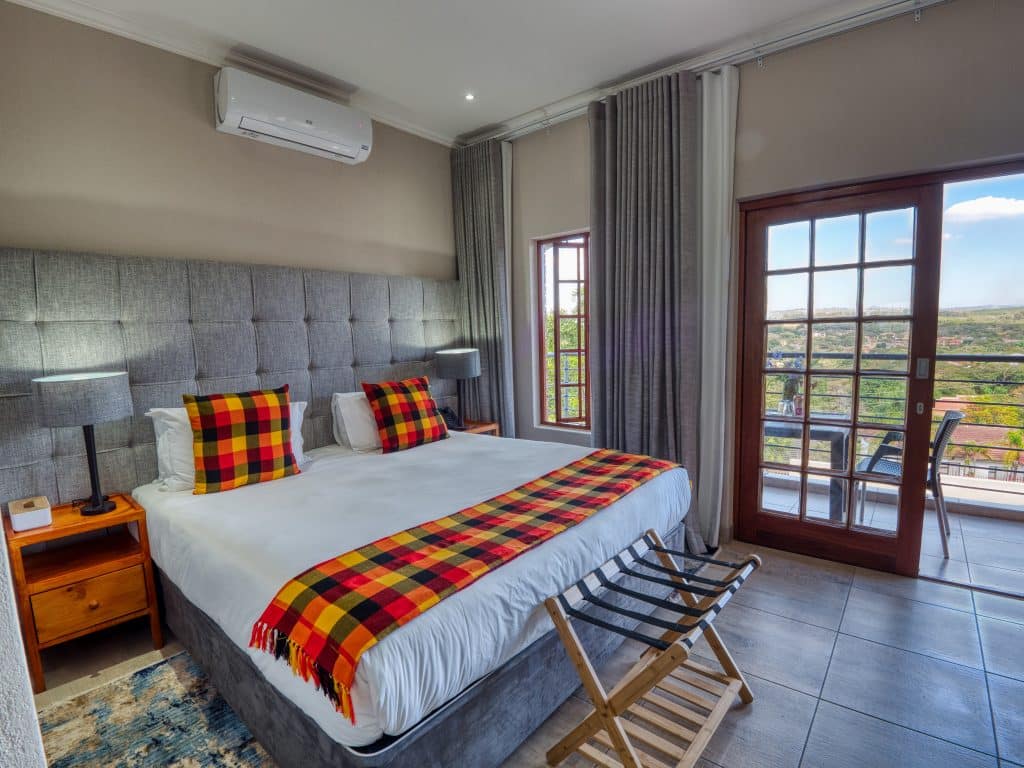 A modern hotel room with a large bed featuring plaid pillows and a matching blanket. Two bedside tables with lamps flank the bed. A window and glass door open to a balcony with views of greenery outside. A luggage rack sits at the foot of the bed.