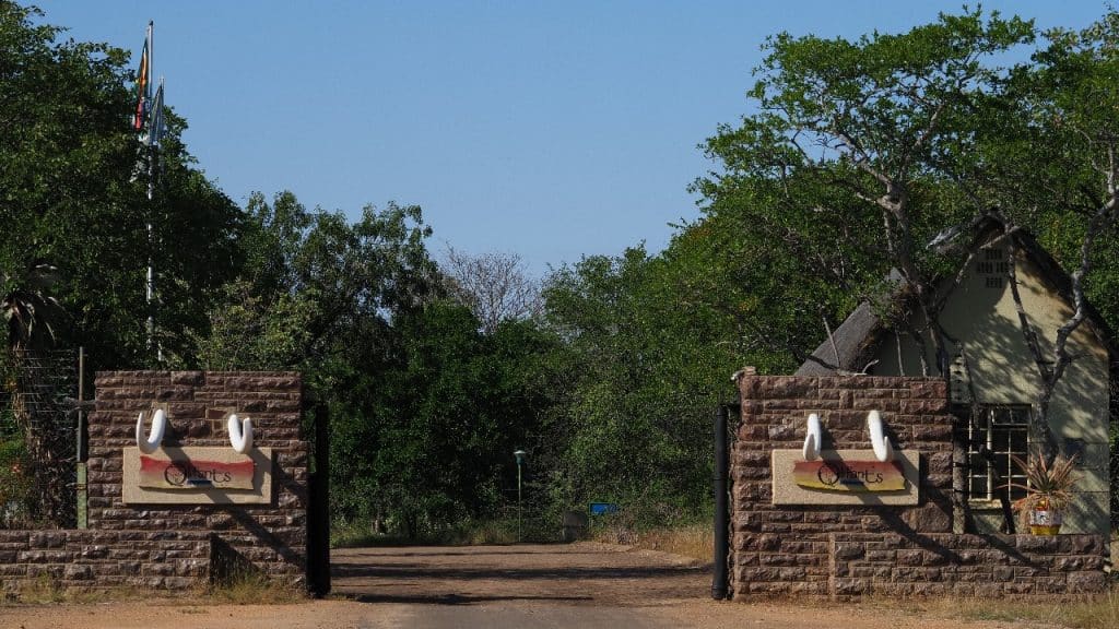 Entrance to a wildlife park with large gates and tusk decorations on either side. A dirt road leads through the gates, surrounded by lush greenery and trees. A small building is visible on the right. Flags are visible in the background.