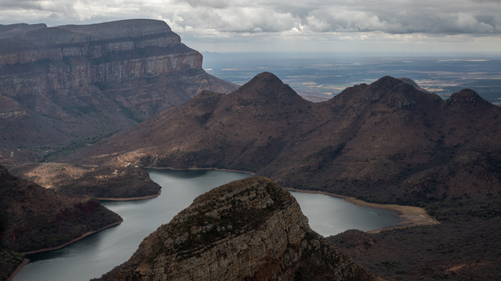 Aerial view of a scenic landscape with rugged mountain ranges and a winding river below. The sky is overcast, casting soft, diffused light over the terrain, highlighting the textures and contours of the rocky formations.