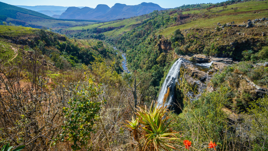 A breathtaking view of the Berlin Falls in South Africa, cascading down a rugged cliff amidst lush greenery. The river winds through a verdant valley, with mountain peaks visible in the distance under a clear blue sky.