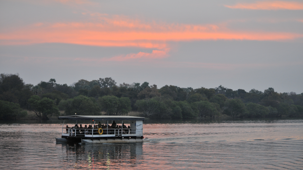 A boat with passengers sails on a calm river during sunset. The sky is painted with shades of orange and pink, and lush green trees line the riverbank, reflecting in the water.